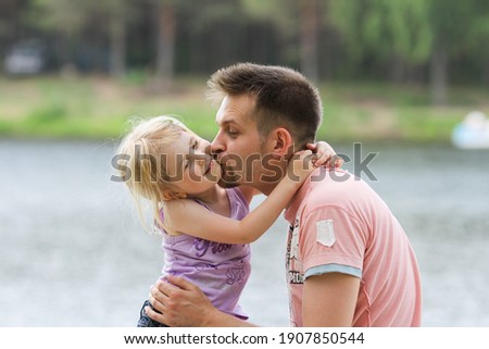 Dad hugging and kissing his little cute daughter on the shore of a lake. Family spending time together in the contry. Father-daughter relationship, father's day, parental care. Selective soft focus.