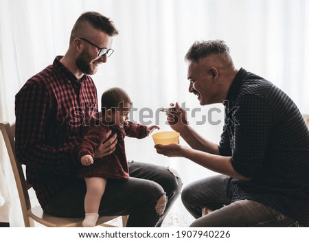 Male gay couple with adopted baby girl at home - Two handsome dads feed the baby girl on kitchen - Lgbt family at home, Diversity concept - Vintage filter Royalty-Free Stock Photo #1907940226