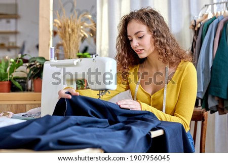 dressmaker tailoring while sitting at sewing machine looking seriously and confident, in light modern atelier room. garment, design, fashion, dressmaking concept Royalty-Free Stock Photo #1907956045