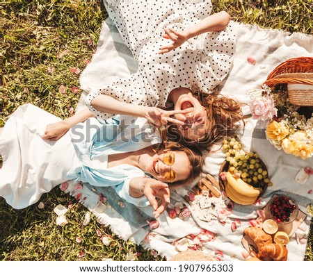 Two young beautiful smiling hipster female in trendy summer sundress and hats.Carefree women making picnic outside.Positive models sitting on plaid on grass, hugging, eating fruits and cheese Royalty-Free Stock Photo #1907965303