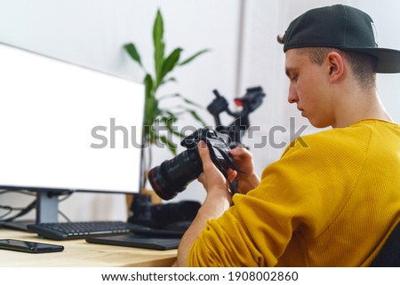 young man photographer, sets up camera, views photos and videos on camera at home. teenager processes photos and videos. Monitor with white screen. Freelancer. Remote work. Retouching training