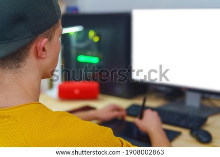 Over-the-shoulder view, young man in baseball cap and yellow sweater, working on pen tablet at computer at home. monitor has white screen. Retouching. Freelancer. Remote work. Selective focus.