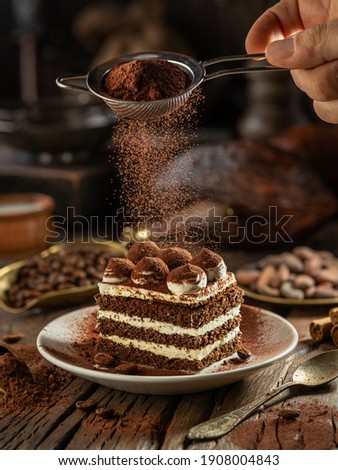 Dusting tiramisu-like cake with cocoa powder. Still life with slice of cake and coffee and cacao beans on wooden table. Royalty-Free Stock Photo #1908004843