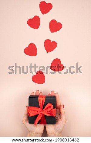 Festive present box with red bow in female hands. Red paper hearts on pink background. High quality photo