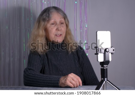 An elderly woman sits at a table in front of a smartphone on a tripod. Senior woman blogger is recording a video that is broadcast online indoors