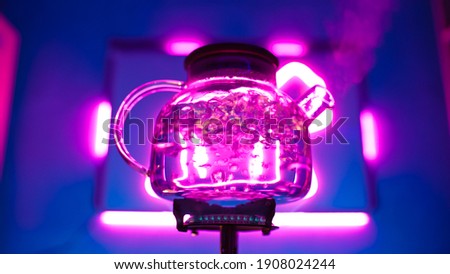 A transparent kettle boils on a gas burner against a neon purple backlight. Blurred bubbles in hot water boiling inside a glass teapot.