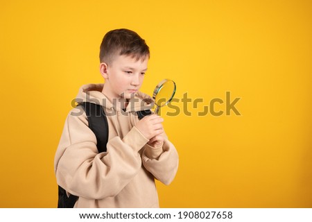Serious boy looking through a magnifying glass, a teenager in search of, looking at something isolated on yellow background. High quality photo