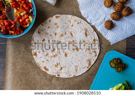 Fresh gluten free tortilla surrounded with chickpea falafel, fresh vegetables chopped salad, jalapenos, mayo and scallions. Healthy and tasty meal preparation for a falafel wrap. Top view.