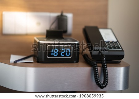 Digital alarm clock at hotel room sitting on bedside table along with the telephone and note pad. Time management concept. Selective focus.
