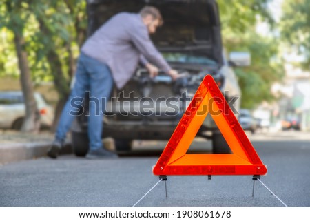 Man fixing car on road. Car Breakdown while driving. Driver looks on breakdown under the hood of car. Red triangular stop sign on road.