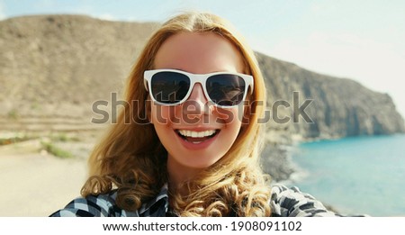 Summer vacation portrait of happy smiling young woman stretching hand for taking selfie on smartphone on the beach on sea and mountain background