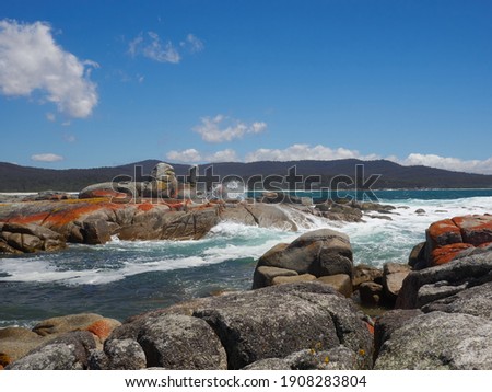 Binalong Bay Tasmania in full sun, rocks along the coastline are red lichen encrusted. The water is a perfect azure blue and the blue sky almost cloudless.         