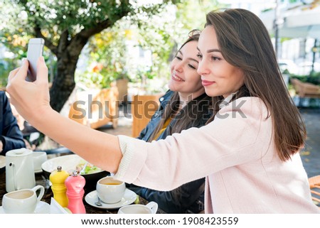 Two young happy brunette women friends making selfie on smartphone during brunch in cafe outside on sunny day