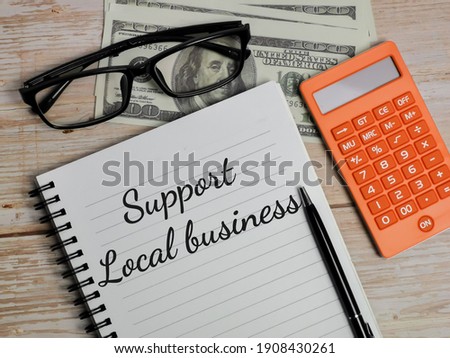 Top view text Support Local business written on notebook with pen,calculator,fake money and eye glasses on wooden table. Finance concept.