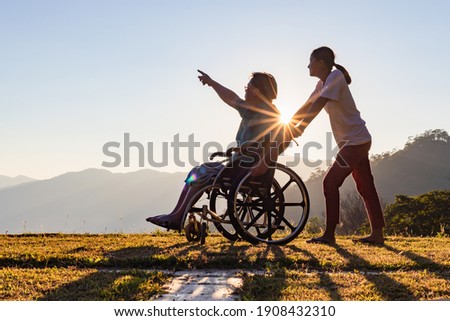 Disabled handicapped young man in wheelchair walking with his care helper in sunset.Silhouette Royalty-Free Stock Photo #1908432310