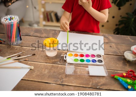 child's hand with a brush and paints draws on white paper on a wooden table with stationery. Creative development of the child