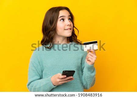 Little caucasian girl isolated on yellow background buying with the mobile with a credit card while thinking