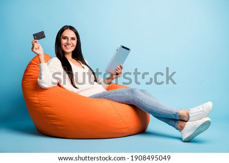 Full length profile photo of attractive cheerful lady sit soft chair hold tablet debit card isolated on blue color background