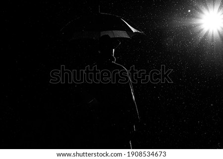 dark silhouette of a man in a raincoat and hat under an umbrella on the street in the rain on the road at night