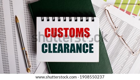 On the desktop are reports, glasses, a pen, a green diary and a white notebook with the words CUSTOMS CLEARANCE. Workplace close-up. Business concept
