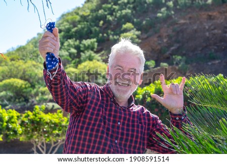 Amused senior man holding a protective coronavirus mask in his hand. He smiles looking at the camera gesturing a positive sign. A joyful elderly person with white hair and beard