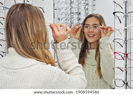 Woman chooses glasses in the store. Blonde in a beige sweater measures glasses near the mirror. Girl on a background of shop windows with different models of glasses. Royalty-Free Stock Photo #1908594586