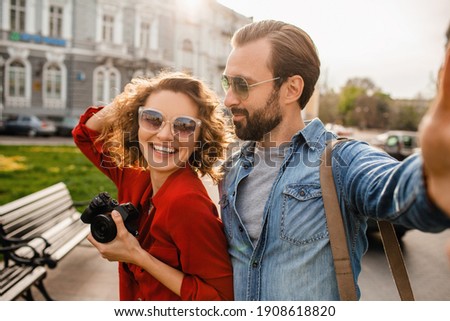 attractive laughing man and woman traveling together, stylish couple in love taking selfie photos on phone on romantic trip, sunny summer city, wearing shirt, sunglasses, travelers having fun