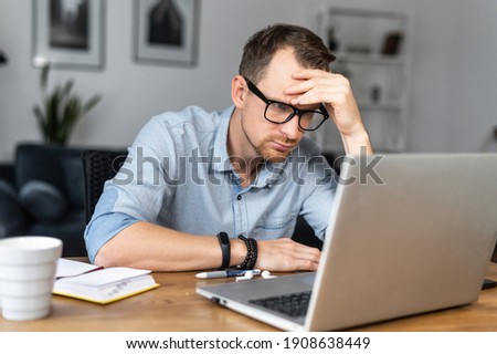 Thoughtful young man in modern glasses and smart casual wear is looking on laptop screen and solving some tasks. A guy works online from home Royalty-Free Stock Photo #1908638449