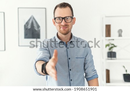 Young pleasant man wearing glasses and smart casual shirt holds out his hand in handshake gesture, greeting, sign of good deal standing in a contemporary office space