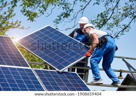 Two young technicians mounting heavy solar photo voltaic panel on tall steel platform on green tree background. Exterior solar panel voltaic system installation, dangerous job concept. Royalty-Free Stock Photo #1908659404