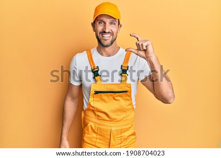 Young handsome man wearing handyman uniform over yellow background smiling and confident gesturing with hand doing small size sign with fingers looking and the camera. measure concept. 
