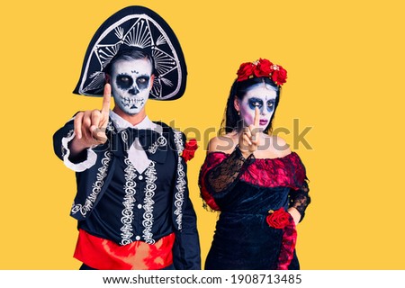 Young couple wearing mexican day of the dead costume over background pointing with finger up and angry expression, showing no gesture 