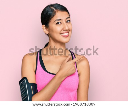 Beautiful asian young sport woman wearing sportswear and arm band cheerful with a smile of face pointing with hand and finger up to the side with happy and natural expression on face 