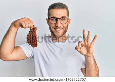 Young caucasian man holding beef steak doing ok sign with fingers, smiling friendly gesturing excellent symbol 