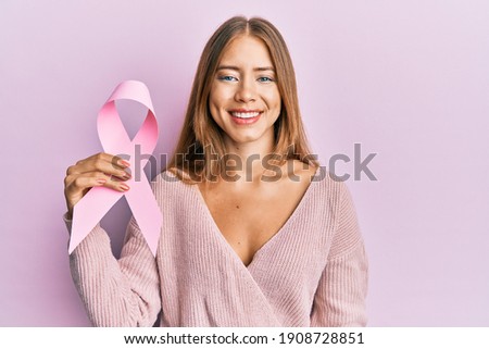 Beautiful young blonde woman holding pink cancer ribbon looking positive and happy standing and smiling with a confident smile showing teeth 
