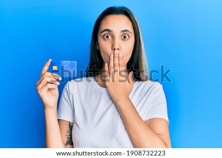Young hispanic woman holding credit card covering mouth with hand, shocked and afraid for mistake. surprised expression 