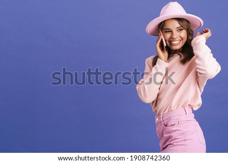 Happy charming girl in hat smiling while talking on cellphone isolated over purple background