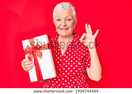 Senior beautiful woman with blue eyes and grey hair holding gift doing ok sign with fingers, smiling friendly gesturing excellent symbol 