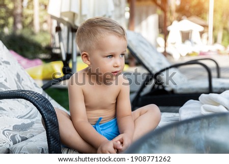 Cute adorable caucasian blond little pensive kid boy enjoy having fun relaxing sitting on sunbed in yard garden near pool sea beach. Child chilling outdoor on sunny day. Toddler sunbathing outside