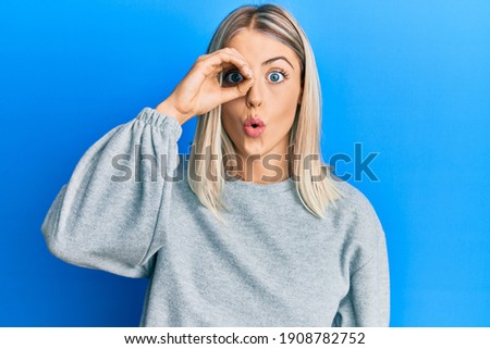 Beautiful blonde woman wearing casual clothes doing ok gesture shocked with surprised face, eye looking through fingers. unbelieving expression. 