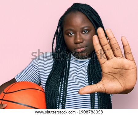 Young black woman with braids holding basketball ball with open hand doing stop sign with serious and confident expression, defense gesture 