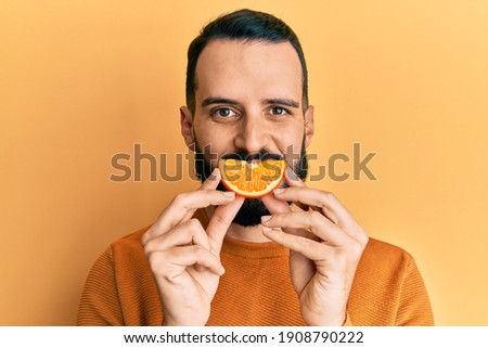 Young man with beard holding orange slice on mouth as funny smile looking positive and happy standing and smiling with a confident smile showing teeth  Royalty-Free Stock Photo #1908790222