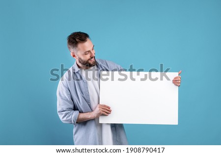 Happy smiling man displaying white blank placard on blue studio background, mockup for logo or design. Positive millennial guy with empty advertising banner, copy space