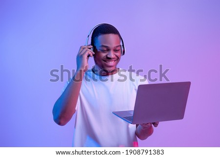 Black Gamer Man In Wireless Headset Playing Video Games On Laptop, Looking At Computer Screen While Standing In Vivid Neon Light Over Purple Studio Background, Guy Enjoying Cyber Gaming, Copy Space