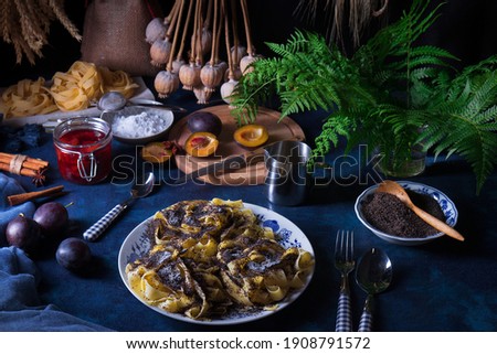 Sweet pasta dessert, noodles with poppy seeds, plum compote from fresh plums, dark background, blue table decorated with fresh flowers. Dry whole poppy plant in background. 