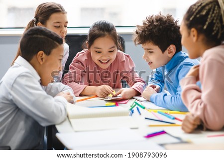 Technology And Pupils Concept. Group of excited multicultural happy junior children sitting at table and using smartphone, playing online mobile games. Modern Device, Gadget Addiction Royalty-Free Stock Photo #1908793096
