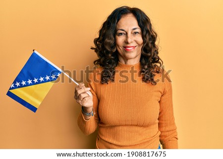Middle age hispanic woman holding bosnia herzegovina flag looking positive and happy standing and smiling with a confident smile showing teeth  Royalty-Free Stock Photo #1908817675