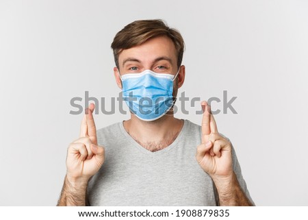 Concept of pandemic, covid-19 and social-distancing. Hopeful worried guy in medical mask, cross fingers for good luck and making wish, standing over white background