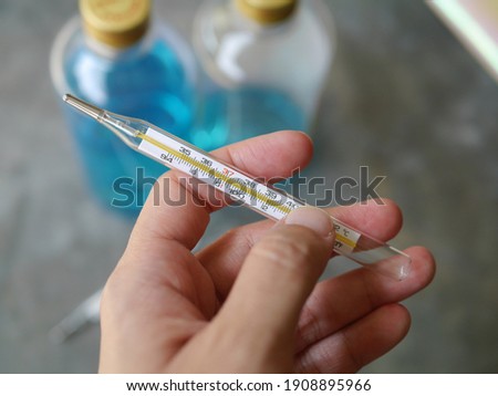Hand held a close-up of a glass thermometer. Behind there is a picture of hand washing alcohol during the Covid-19 outbreak.