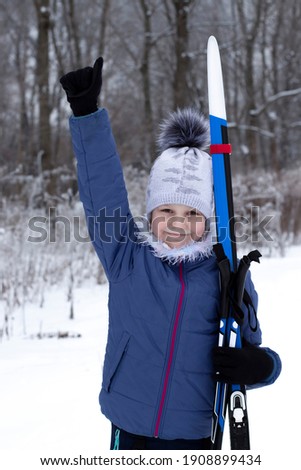A nine-year-old young athlete pulls her hand up. In the other hand he holds skis and ski poles. Winter sports. Classic cross-country skiing.